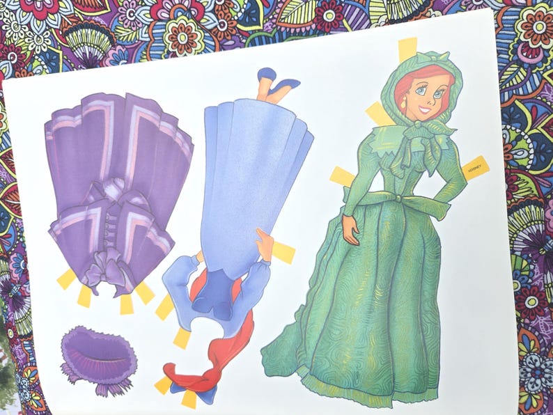 May include: A paper doll set with a green dress, a purple jacket, and a blue dress with a red scarf. The paper doll has a green dress and bonnet. The set is on a white background.