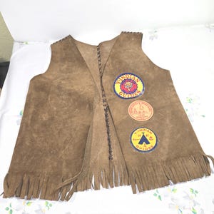 May include: Brown suede vest with fringe along the bottom edge. The vest has three patches with text and graphics. The vest is open in the front and laced together with brown thread. The vest is likely vintage.