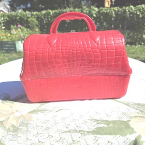 May include: A bright red vintage handbag with a textured, crocodile-like pattern. The bag has a handle and a clasp closure. The word "PEERLESS" is embossed on the handle. The bag is sitting on a floral patterned tablecloth.