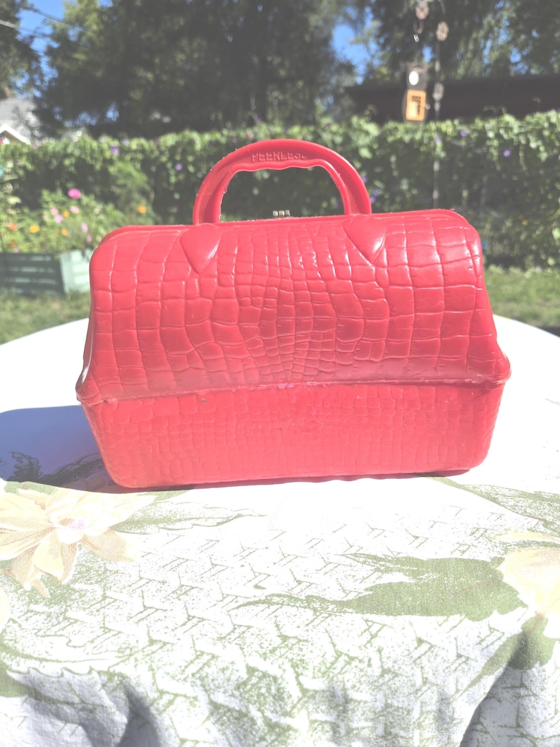 May include: A bright red vintage handbag with a textured, crocodile-like pattern. The bag has a handle and a clasp closure. The word "PEERLESS" is embossed on the handle. The bag is sitting on a floral patterned tablecloth.