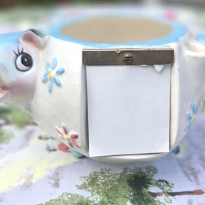 May include: A ceramic pig-shaped planter with a light blue and white color scheme. The pig has a smiling face, blue flower accents, and a small rectangular opening on its side. The planter is on a patterned surface.