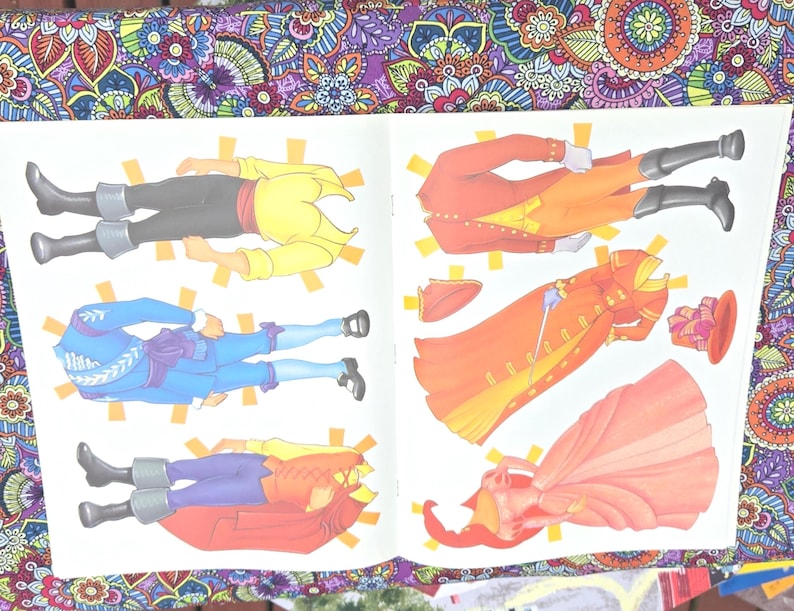 May include: An open book displaying paper dolls of historical figures. The dolls are dressed in outfits of yellow, blue, red, and orange. The paper dolls are cut out and ready to be played with.