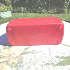 May include: A red rectangular box with a textured surface and rounded corners. The box has four small, rounded feet. The box is sitting on a floral patterned tablecloth.