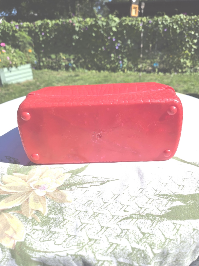 May include: A red rectangular box with a textured surface and rounded corners. The box has four small, rounded feet. The box is sitting on a floral patterned tablecloth.