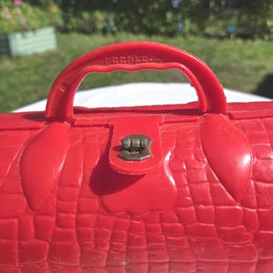 May include: A red vintage plastic purse with a textured crocodile skin pattern. The purse has a handle with the word "PEERLESS" embossed on it, and a metal clasp closure. The purse is a bright, solid red color.