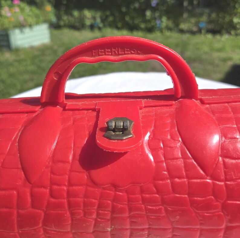 May include: A red vintage plastic purse with a textured crocodile skin pattern. The purse has a handle with the word "PEERLESS" embossed on it, and a metal clasp closure. The purse is a bright, solid red color.