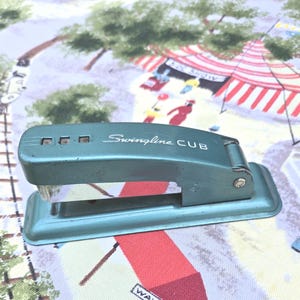 May include: Vintage teal Swingline CUB stapler. The stapler has the words "Swingline CUB" printed on the side. The stapler is made of metal and has a rectangular base. The background is a patterned fabric with circus and tree designs.
