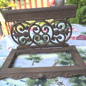 May include: An antique, cast iron plant stand with a rectangular top and ornate scrollwork. The stand features three circular decorative elements and a rectangular base. The metal has a weathered, brown finish, suggesting age and outdoor use.