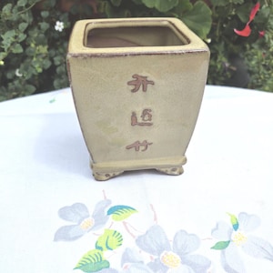 May include: A square, light green ceramic planter with a slightly textured surface. The planter has a dark brown rim and base, with Chinese characters etched on the front. The planter is sitting on a white floral patterned tablecloth.