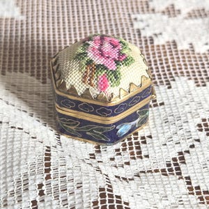 May include: A small hexagonal trinket box with a hinged lid featuring a needlepoint rose design in pink and green on a cream background. The sides are made of dark blue enamel with gold-tone metal trim and painted green leaf and blue flower details.