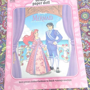 May include: A pink "Deluxe Paper Doll" book featuring Disney's "The Little Mermaid." The cover shows Ariel and Prince Eric holding hands, with a seaside background. The book includes precut clothes and a storage envelope.