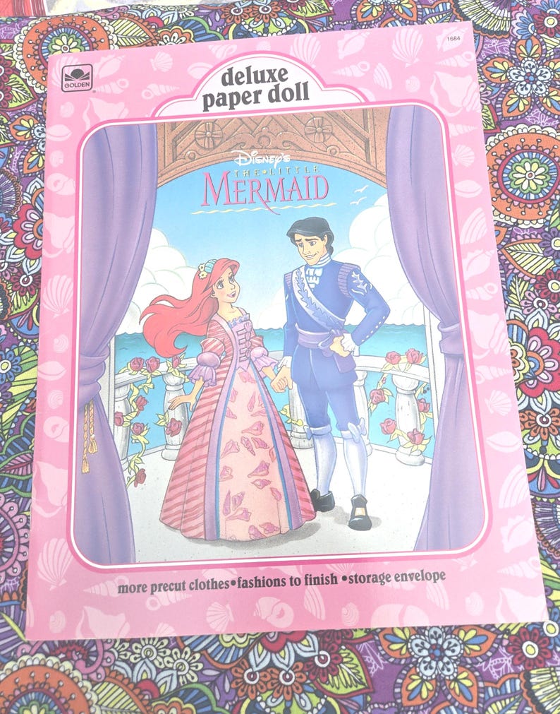 May include: A pink "Deluxe Paper Doll" book featuring Disney's "The Little Mermaid." The cover shows Ariel and Prince Eric holding hands, with a seaside background. The book includes precut clothes and a storage envelope.