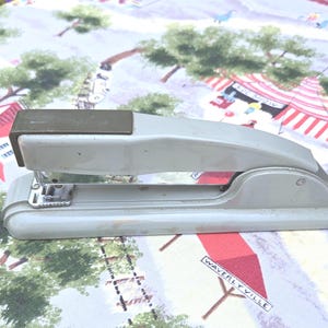 May include: A vintage gray stapler with a silver metal staple holder. The stapler is resting on a colorful patterned fabric with a town scene. The stapler is a classic office supply item.