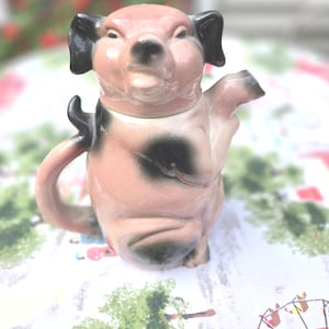 May include: A ceramic teapot shaped like a pig, with a pink body, black ears, and a black spot. The teapot has a handle and a spout, and is sitting on a patterned tablecloth.