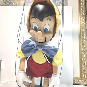 May include: A vintage Pinocchio marionette puppet. The puppet has a yellow hat, black hair, and blue eyes. It wears a yellow shirt, red overalls, a blue bow tie, and white gloves. The puppet is attached to a wooden control.