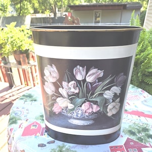 May include: Black metal wastebasket with a white band and a floral design. The design features a bouquet of pink, white, and purple tulips in a blue and white patterned bowl against a black background. The wastebasket is cylindrical.