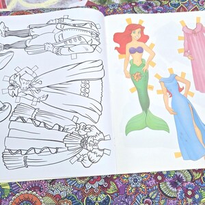 May include: An open paper doll book with illustrations of paper dolls and their outfits. The left page shows black and white line drawings of a man and two dresses. The right page shows a mermaid and two dresses in colour.