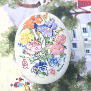 May include: Oval ceramic trinket box with a floral design. The lid features a bouquet of colorful flowers, including roses, irises, and pansies, in shades of pink, orange, yellow, and blue. The artist's signature "Suzanne Clee" is visible.