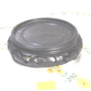May include: A dark brown, circular wooden display stand featuring a flat top surface with a recessed center and a carved, openwork base with scalloped edges. The stand has a smooth, matte finish and is designed to support decorative objects.