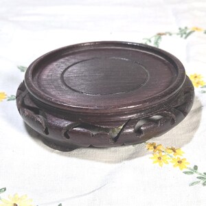 May include: A circular dark brown wooden display stand featuring a smooth, flat top surface with a recessed center and a carved, scalloped base with openwork detailing along the sides, providing a stable platform for small decorative objects.