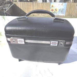 May include: Black vintage American Tourister suitcase with a top handle and silver-toned accents. The suitcase has a rectangular shape with rounded edges and small feet. The American Tourister logo is visible on the front.