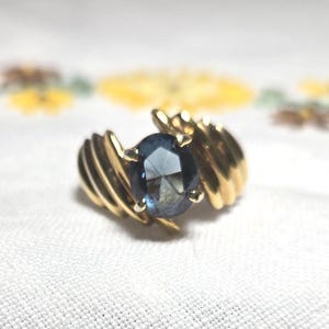 May include: A gold ring featuring a dark blue oval gemstone. The ring has a ribbed design on the sides, and the gemstone is set in a prong setting. The ring is on a white surface with a floral pattern in the background.