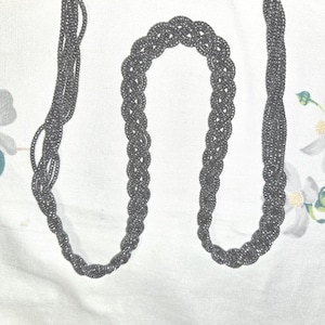 May include: A dark gray beaded necklace with a braided central section and multiple strands. The necklace is displayed on a white surface with floral accents, showcasing its intricate design and texture.