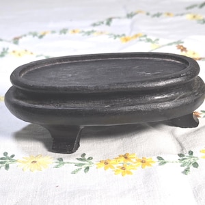 May include: An oval-shaped black wooden display stand featuring a raised rim and a smooth, flat top surface. The base is supported by four short, sturdy feet, providing a stable platform for elevating small decorative objects or collectibles.