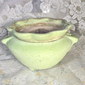 May include: A light green ceramic planter with a speckled finish and a scalloped rim. The bowl-shaped vessel features two small side handles and rests on three short feet, with a weathered, brownish interior showing signs of past use.
