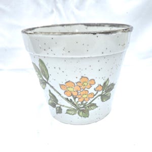 May include: A ceramic flower pot with a speckled white finish and a brown rim. The pot features a floral design with orange flowers and green leaves. The pot is designed for planting flowers or herbs.
