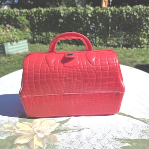 May include: A vintage red plastic handbag with a textured, crocodile-like pattern. The bag has a handle, a metal clasp, and the word "Peerless" embossed on the handle. The bag is sitting on a floral tablecloth.
