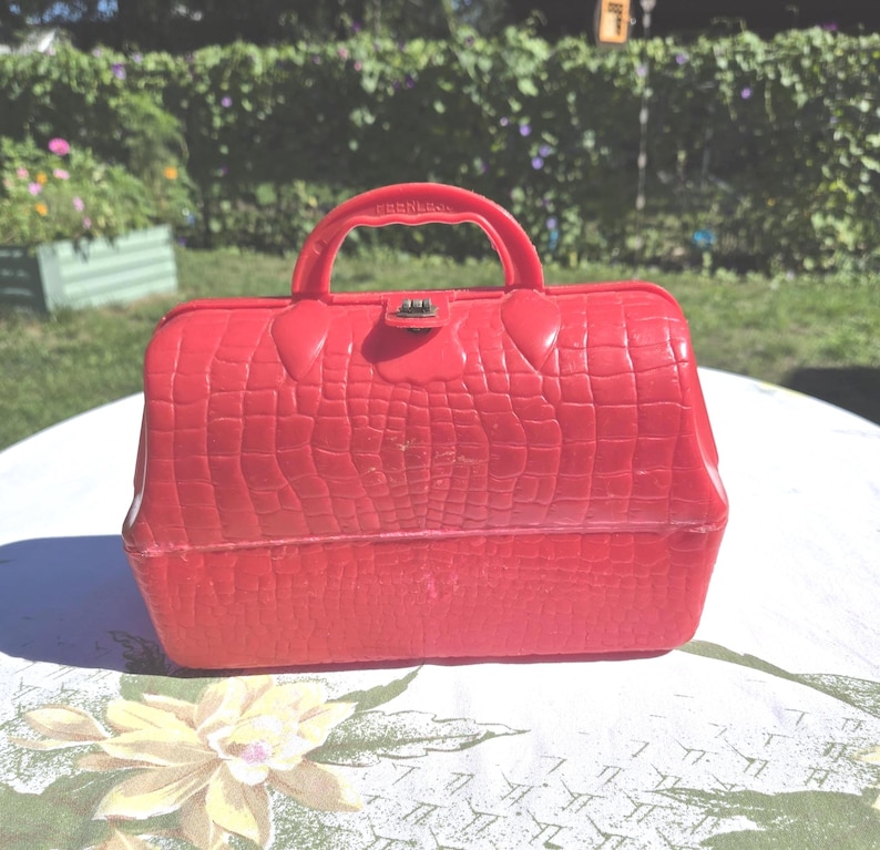 May include: A vintage red plastic handbag with a textured, crocodile-like pattern. The bag has a handle, a metal clasp, and the word "Peerless" embossed on the handle. The bag is sitting on a floral tablecloth.