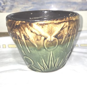 May include: A ceramic bowl with a dark brown rim and a green base. The bowl features a textured design with leaf and fruit motifs in shades of brown and tan. The bowl is sitting on a white surface.