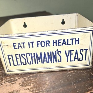 May include: Vintage rectangular metal container with the text "EAT IT FOR HEALTH FLEISCHMANN'S YEAST" in blue lettering on a cream background. The container has two holes for hanging and is placed on a wooden surface.