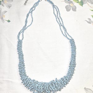 May include: A light blue beaded necklace with a multi-strand design. The necklace features a cluster of beads at the center, with a delicate chain and clasp. The jewelry is displayed on a white floral patterned background.