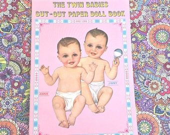 May include: A pink paper doll book titled "The Twin Babies Cut-Out Paper Doll Book." The cover features illustrations of two babies, "Annie" and "Jimmy." The book is set against a colorful floral patterned background.