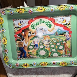 May include: A vintage green metal tray with a colourful Cabbage Patch Kids design. The tray has a floral border with yellow, pink, and blue flowers. The central image depicts Cabbage Patch Kids in a field with a tree and a red banner.