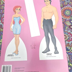 May include: A pink paper doll book featuring Ariel and Eric from Disney's The Little Mermaid. Ariel is in a blue outfit, and Eric is shirtless with black trousers. The book has the text "ARIEL" and "ERIC".