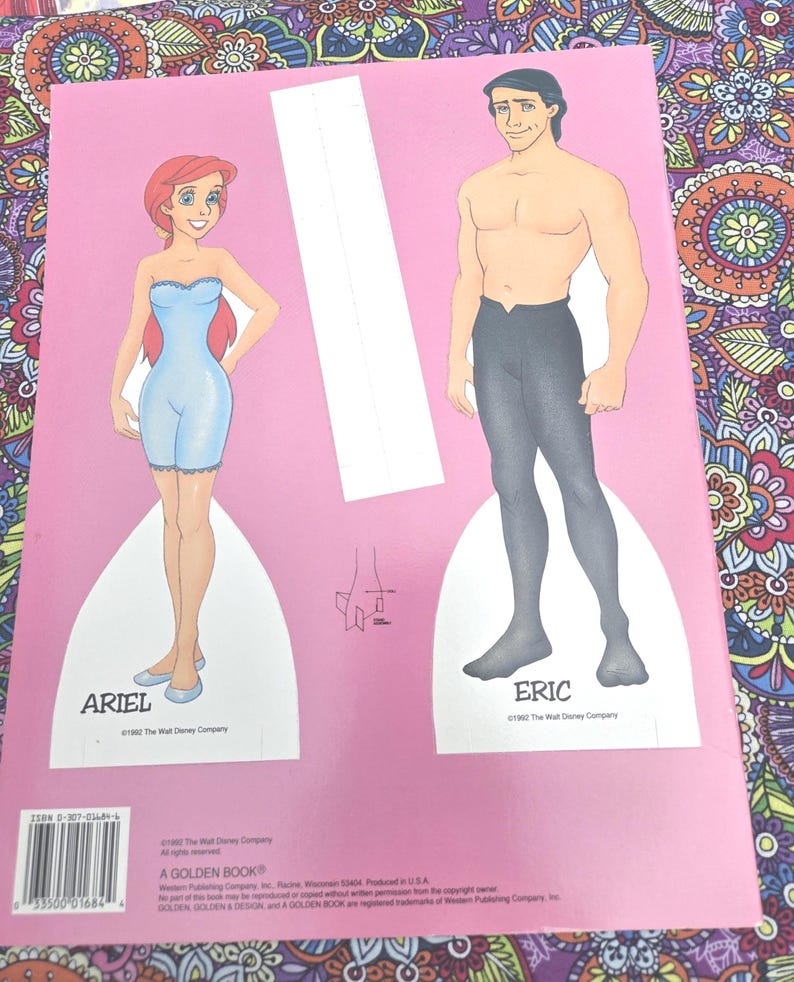 May include: A pink paper doll book featuring Ariel and Eric from Disney's The Little Mermaid. Ariel is in a blue outfit, and Eric is shirtless with black trousers. The book has the text "ARIEL" and "ERIC".