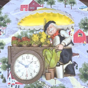 May include: A decorative wall clock featuring a smiling elderly man with a red scarf, black hat, and vest, pushing a cart of flowers under a yellow umbrella. The clock face is white with black numbers and hands. The background depicts a rural scene.