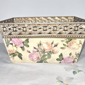 May include: Rectangular decorative basket with a floral rose design. The basket has a cream-colored background with pink and peach roses and green leaves. The top edge has a gold-colored filigree design.