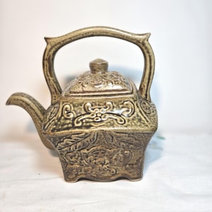 May include: A ceramic teapot with a detailed floral and scroll design. The teapot is a muted olive green color with a matching lid and a curved handle. The spout is short and angled. The teapot sits on four small feet.