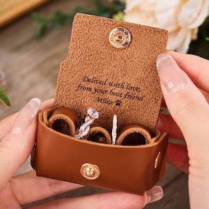 Puede incluir: Un joyero de cuero marrón con cierre de botón a presión, que contiene dos anillos. La caja está abierta, revelando los anillos y el texto "Delivered with love, from your best friend Miles".