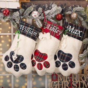 Peut inclure: Trois bas de Noël blancs avec des motifs de pattes et des accents écossais. Chaque bas est personnalisé avec un nom : William, Ashley et Max. Les bas sont décorés d'ornements en forme d'os et de poisson. Les mots "MERRY CHRISTMAS" sont visibles.