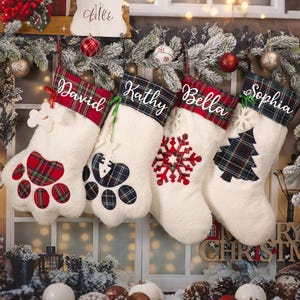 May include: Four white Christmas stockings with personalized names: David, Kathy, Bella, and Sophia. Each stocking has a plaid cuff and a unique design: paw prints, a snowflake, and a Christmas tree. Festive holiday decor.