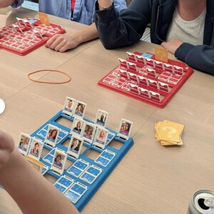 May include: Two Guess Who board games with red and blue plastic frames featuring rows of flip-up character cards. Each game includes a yellow card with a question mark symbol. The boards are set on a flat surface with scattered game cards.
