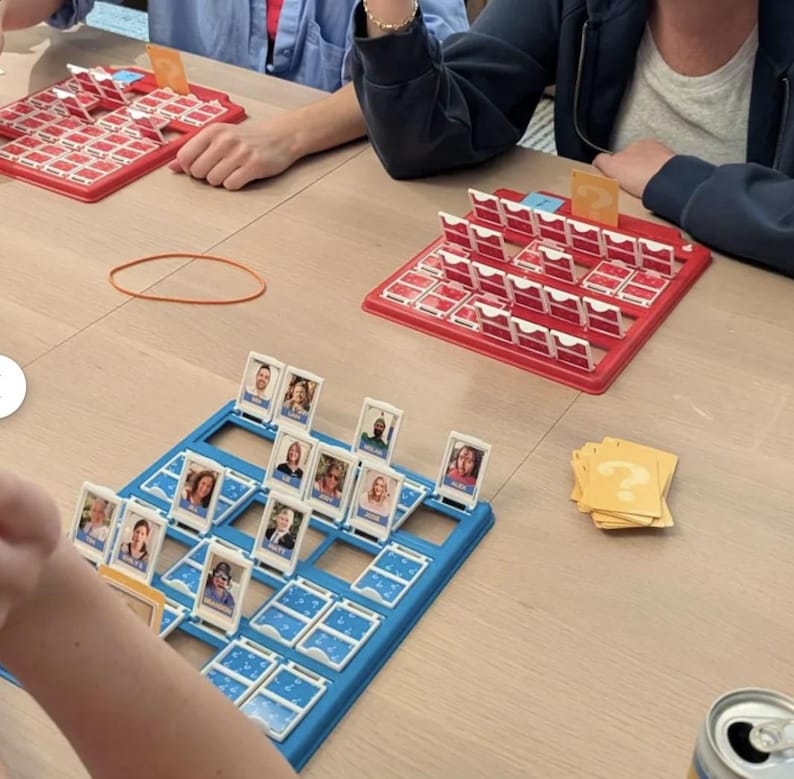 May include: Two Guess Who board games with red and blue plastic frames featuring rows of flip-up character cards. Each game includes a yellow card with a question mark symbol. The boards are set on a flat surface with scattered game cards.