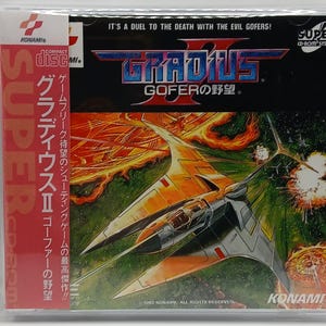 May include: A Super CD-ROM System game case for Gradius II: Gofer's Ambition. The cover art features a futuristic spaceship in flight, with a fiery orange and green background. The game title is prominently displayed in blue and red. The case is in a clear plastic sleeve.