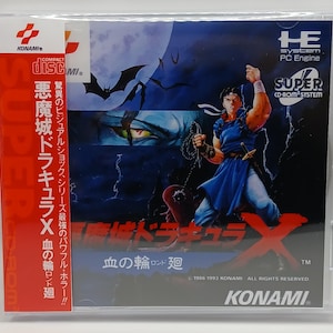 May include: A video game case for "Castlevania: Rondo of Blood" featuring a muscular character in blue attire, holding a weapon. The cover art includes the game title in Japanese characters, a bat silhouette, and the Konami logo. The case is in a plastic sleeve.