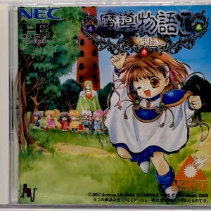 May include: A PC Engine video game case for Madou Monogatari I, featuring a colorful anime-style illustration of a character in a white and blue outfit running through a grassy field with a tall tower and smaller figures in the background.
